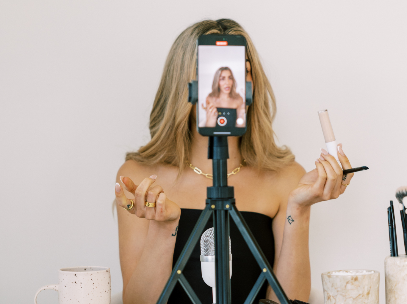 A woman is seated behind a tripod-mounted smartphone camera, recording herself while demonstrating a makeup product. The phone is centered and captures her face clearly on-screen. She’s holding a makeup item (possibly concealer or foundation) in one hand and a brush or applicator in the other.